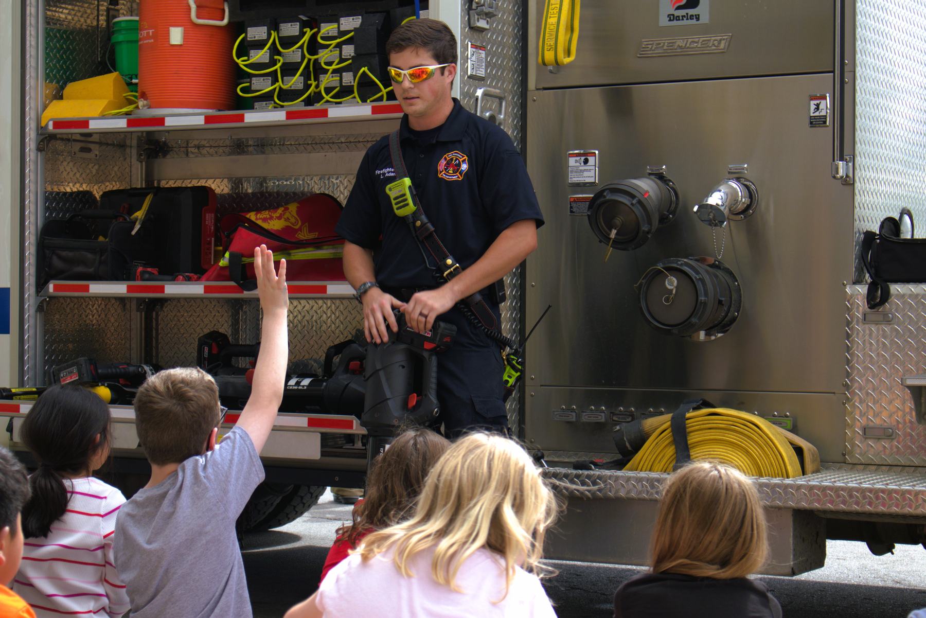 Students raising hands as a firefighter shows equipment on the engine